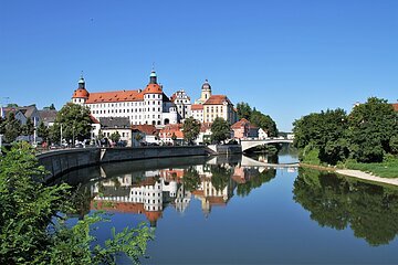 Schloss Neuburg und die Donau, die sich rechts am Schloss vorbeischlängelt. Links ist die Promenade und links das begrünte Ufer zu sehen.