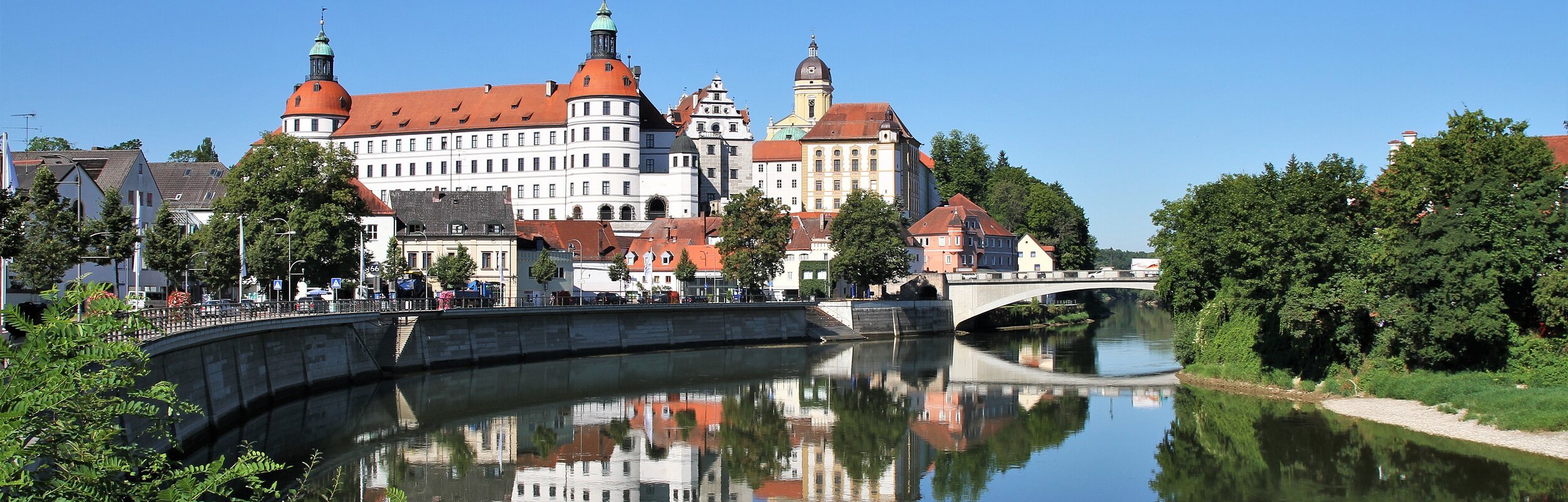 Schloss Neuburg und die Donau, die sich rechts am Schloss vorbeischlängelt. Links ist die Promenade und links das begrünte Ufer zu sehen.