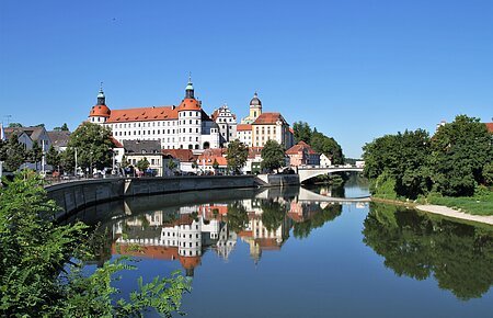 Schloss Neuburg und die Donau, die sich rechts am Schloss vorbeischlängelt. Links ist die Promenade und links das begrünte Ufer zu sehen.