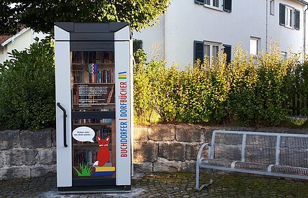 Blick auf den öffentlich zugänglichen Bücherschrank in der Hauptstraße in Buchdorf mit rechts daneben stehender Sitzgelegenheit.