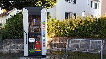 Blick auf den öffentlich zugänglichen Bücherschrank in der Hauptstraße in Buchdorf mit rechts daneben stehender Sitzgelegenheit.