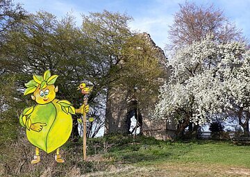 Blick auf eine zwischen Bäumen versteckte Ruine. Links sieht man das grüne, gemalte Waldgeistlein.