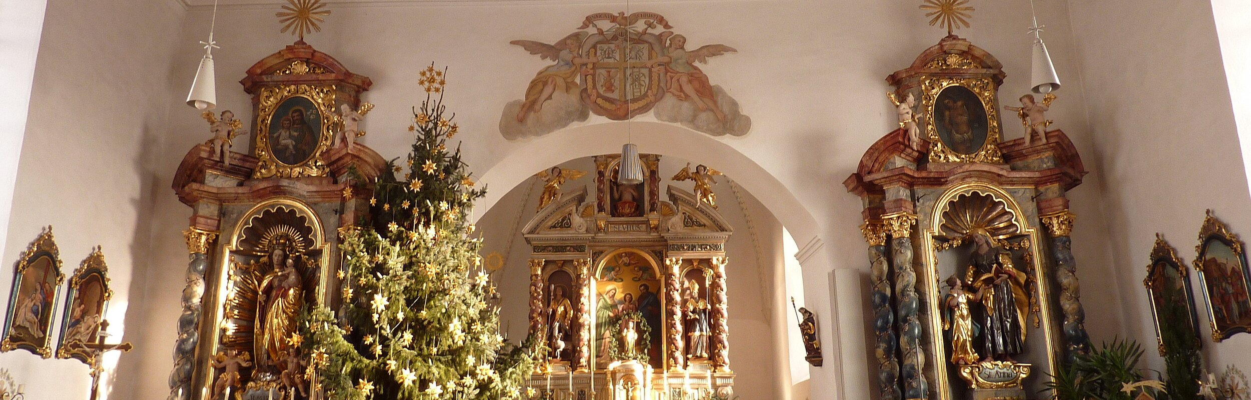 Blick auf den Hauptaltar und die beiden Seitenaltäre zur Weihnachtszeit mit geschmücktem Weihnachtsbaum. Vor dem rechten Seitenaltar ist die Krippe aufgebaut.