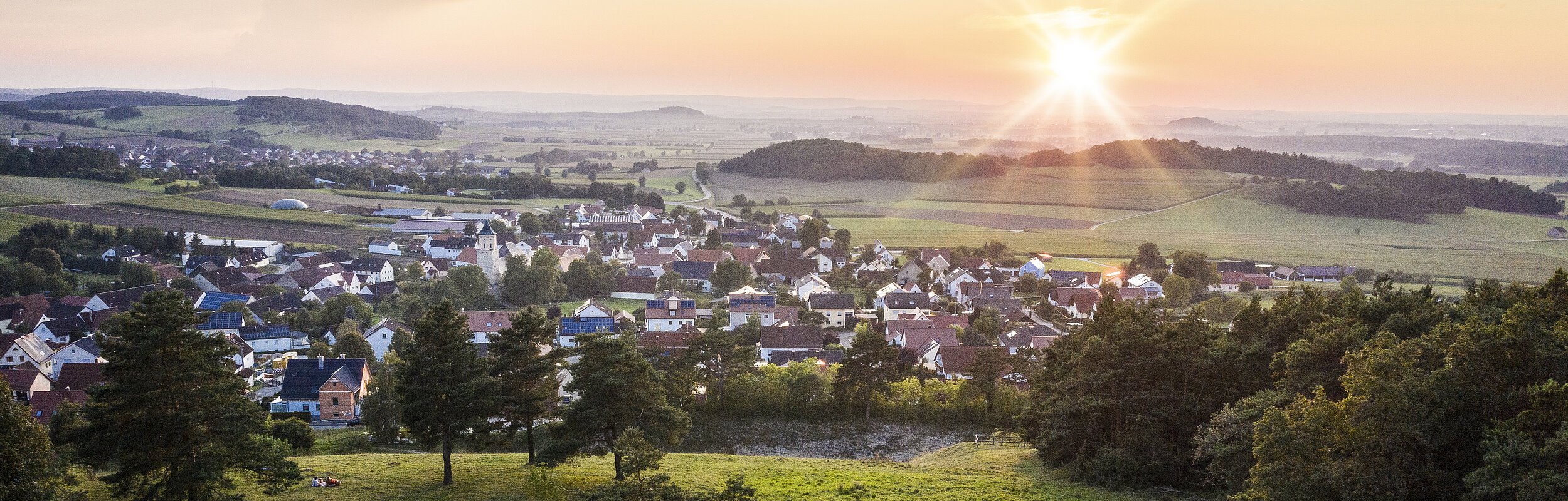 Der Blick von oben über grüne Wiesen hinab auf die Stadt Monheim. Die Sonne strahlt am Horizont und ist fast untergegangen.