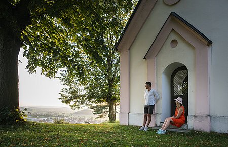 Ein Mann im weißen Shirt lehnt an der Wand einer Kapelle an, eine Frau im roten Kleid und Strohhut sitzt auf der Stufe. Beide genießen den Sonnenuntergang. Links neben der Kapelle steht ein großer Baum mit grünem Blätterdach, dass den Beiden Schatten spendet.