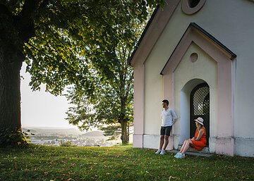 Wanderer auf der Monheimer Alb Ein Mann im weißen Shirt lehnt an der Wand einer Kapelle an, eine Frau im roten Kleid und Strohhut sitzt auf der Stufe. Beide genießen den Sonnenuntergang. Links neben der Kapelle steht ein großer Baum mit grünem Blätterdach, dass den Beiden Schatten spendet.