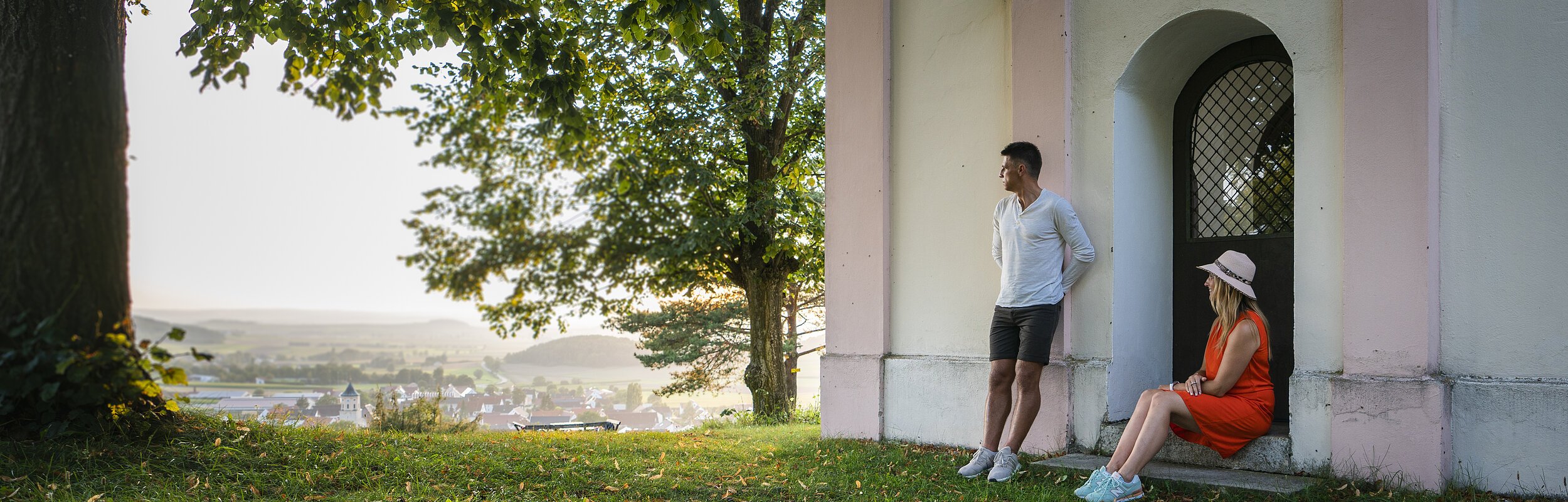 Ein Mann im weißen Shirt lehnt an der Wand einer Kapelle an, eine Frau im roten Kleid und Strohhut sitzt auf der Stufe. Beide genießen den Sonnenuntergang. Links neben der Kapelle steht ein großer Baum mit grünem Blätterdach, dass den Beiden Schatten spendet.