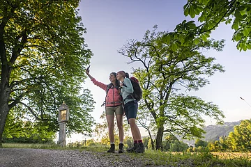 Zwei Frauen in rosafarbenen und hellblauem Shirt, sowie kurzen Hosen stehen eng beieinander am Frauenberg und die Frau in rosa, hält ihr Handy für ein gemeinsames Selfie in die Höhe. Ein großer Baum, sowie ein Marterl sind vor den Zweien zu erkennen. Im Hintergrund der zwei Frauen sind vereinzelt Bäume zu sehen , aber auch ein wolkenloser Himmel.