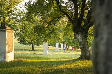 Eine Wandergruppe mit roten und blauen Shirts ist in der Ferne zu erahnen. Sie wandern inmitten grüner Wiesen, umgeben von Bäumen mit grünem Blätterdach. Rechts ist ein gemauertes Denkmal zu sehen.