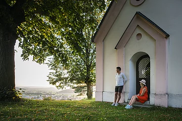 Ein Mann im weißen Shirt lehnt an der Wand einer Kapelle an, eine Frau im roten Kleid und Strohhut sitzt auf der Stufe. Beide genießen den Sonnenuntergang. Links neben der Kapelle steht ein großer Baum mit grünem Blätterdach, dass den Beiden Schatten spendet.