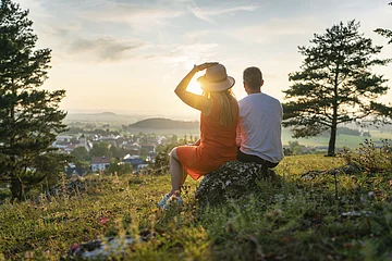 Ein Pärchen sitzt oberhalb von Monheim auf einem Stein umgeben von grüner Wiese und eingerahmt von zwei Bäumen. Die beiden genießen den Sonnenuntergang und den schönen Blick auf Monheim.