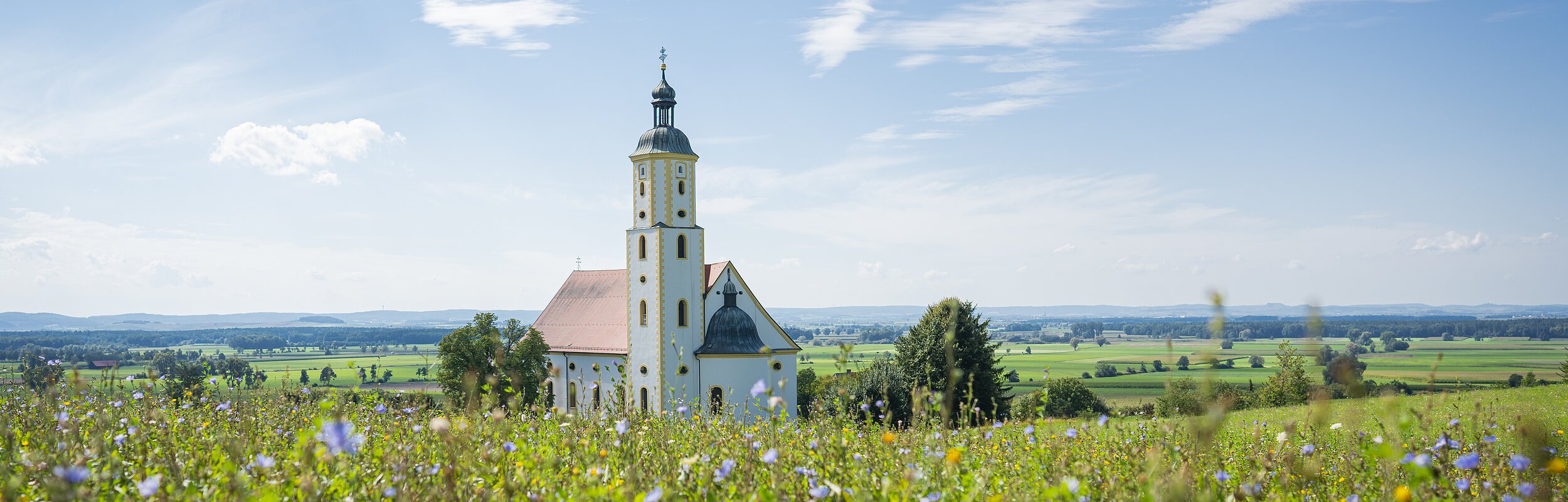 Die Wallfahrtskirche Maria Brünnlein ist von weiten zu sehen. Sie liegt idyllisch inmitten grüner Wiesen. Der Himmel strahlt blau, nur wenige Wolken sind zu sehen.
