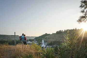 Zwei Wandernde stehen neben dem Kreuz auf dem Pfünzer Kirchberg und schauen über Pfünz zum Osterberg mit seiner Wacholderheide. Über dem Osterberg geht gerade die Sonne unter.