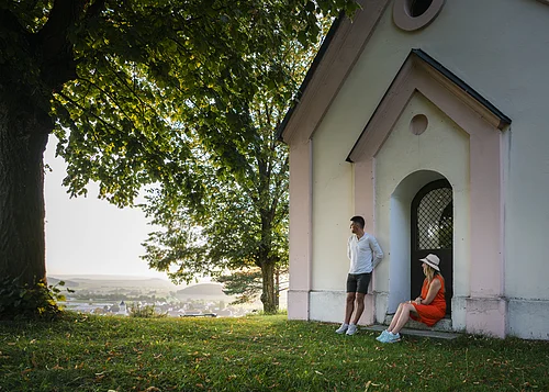 Ein Mann im weißen Shirt lehnt an der Wand einer Kapelle an, eine Frau im roten Kleid und Strohhut sitzt auf der Stufe. Beide genießen den Sonnenuntergang. Links neben der Kapelle steht ein großer Baum mit grünem Blätterdach, dass den Beiden Schatten spendet.