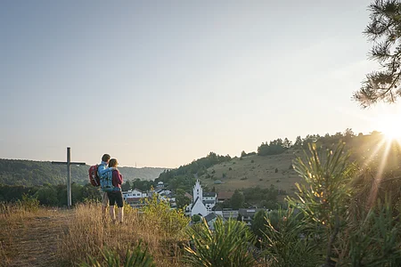 Zwei Wandernde stehen neben dem Kreuz auf dem Pfünzer Kirchberg und schauen über Pfünz zum Osterberg mit seiner Wacholderheide. Über dem Osterberg geht gerade die Sonne unter.