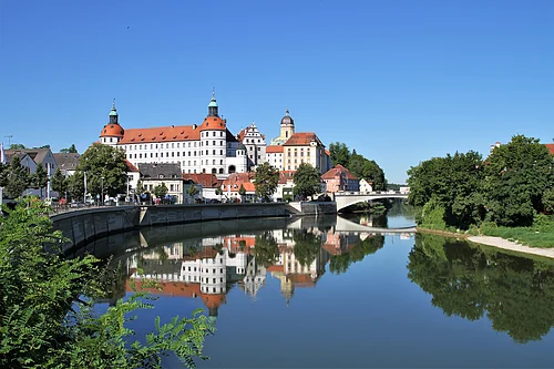 Schloss Neuburg und die Donau, die sich rechts am Schloss vorbeischlängelt. Links ist die Promenade und links das begrünte Ufer zu sehen.