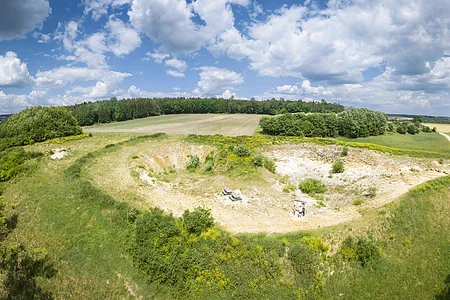 In einer flachen Grube stehen vereinzelt Büsche, während die Hänge aus bloßem Gestein bestehen. Um die Grube herum befinden sich Wälder, Äcker und Wiesen.