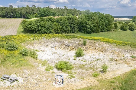 In einer Grube mit vereinzelten Büschen und bloßem Gestein an den Hängen steht eine Sitzbank und eine Informationstafel.
Oberhalb der Grube befinden sich Äcker und Wald.