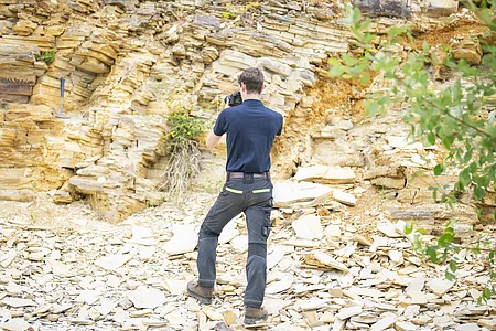 Ein Mann fotografiert mit einer Kamera eine Felswand aus plattigem Kalkgestein. Der Boden ist ebenfalls mit Bruchstücken von Kalkgesteinsplatten bedeckt.