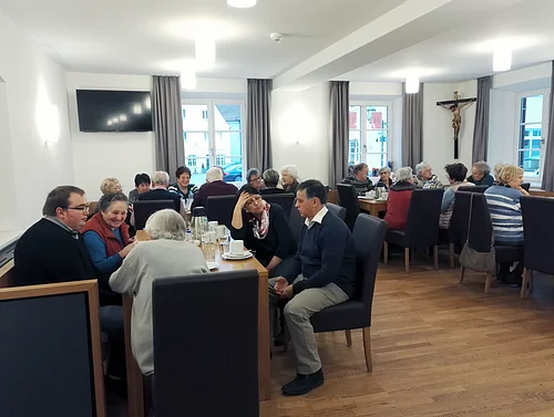 Der Bürgertreff "ZAMSEI" in Monheim: Viele Menschen sitzen bei Kaffee oder Tee in einem Raum an Tischen und unterhalten sich gesellig. Im Hintergrund sind drei Fenster mit Vorhängen und es hängt ein Jesus-Kreuz und ein Fernseher an der Wand.