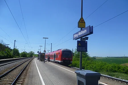 Der Bahnhof Otting-Weilheim mit rotem Zug auf dem Gleis, blauer Himmel, grüne Landschaft im Hintergrund.