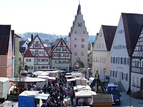 Josefsmarkt in Monheim in der Altstadt: auf dem Marktplatz entlang Fachwerkhäuser, ein Turm mit Uhr und Menschenmengen zwischen vielen Marktständen bei Tageslicht.