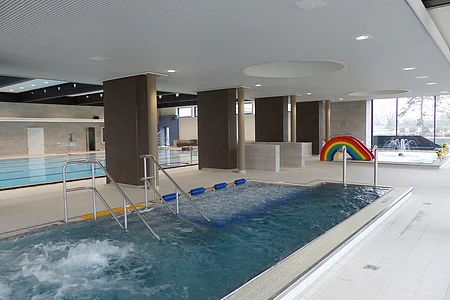 Innenansicht eines Schwimmbads mit mehreren Becken, Rutsche in Regenbogenfarben und Wasserspielen.