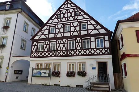 Tourist-Information der Stadt Monheim/Monheimer Alb: ein Fachwerkhaus mit drei Stockwerken, weißen Wänden und braunen Balken. Davor steht eine Sitzbank und ein Schaukasten auf einem gepflasterten Platz. Links davon ein weißes dreistöckiges Gebäude. Rechts davon ein gelbes zweistöckiges Gebäude mit roten Fensterläden.