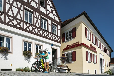 Tourist-Information der Stadt Monheim/Monheimer Alb: ein Fachwerkhaus mit drei Stockwerken, weißen Wänden und braunen Balken. Rechts davon ein gelbes zweistöckiges Gebäude mit roten Fensterläden. Zwei Personen mit Fahrrädern stehen davor und schauen eine Broschüre an.
