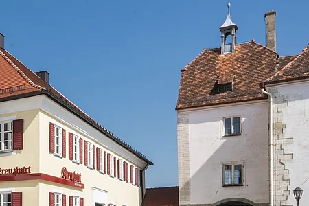Tourist-Information der Stadt Monheim/Monheimer Alb: ein Fachwerkhaus mit drei Stockwerken, weißen Wänden und braunen Balken. Davor steht ein Schaukasten und zwei Radfahrer fahren auf gepflasterter Straße vorbei. Rechts davon ein gelbes zweistöckiges Gebäude mit roten Fensterläden.