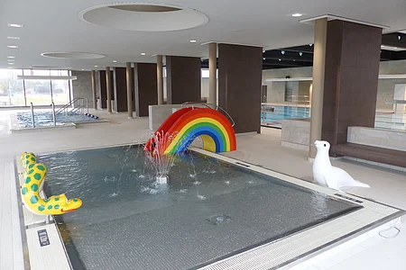 Innenansicht eines Schwimmbads, dem Jurabad Monheim, mit Kinderbecken, Regenbogenrutsche und Wasserspielzeugen. Im Hintergrund ein weiteres Schwimmbecken und ein großes offenes Fenster.