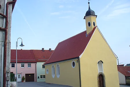 Gelbe Kapelle, die Peterskapelle in Monheim mit rotem Dach und einem Turm, umgeben von Wohnhäusern mit roten Dächern. Im Hintergrund blauer Himmel.