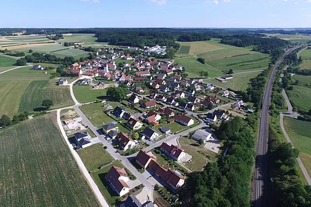 Luftaufnahme des Dorfes Weilheim, mit vielen Häusern, umgeben von Feldern und Wäldern, neben einer Bahnstrecke.