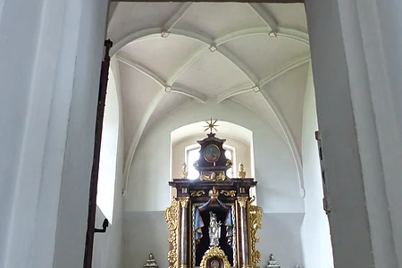 Ein Blick durch eine Tür zu einem Kirchenaltar in der Walburgakapelle in Monheim, mit goldenen Verzierungen und einer Statue, der heiligen Walburga. Der Kirchenaltar ist umgeben von Kerzen und Blumen, in einem gewölbten Raum mit einer verzierten Decke.