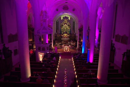 Innenansicht einer Kirche mit violetter Beleuchtung, Kerzen entlang des Gangs und Menschen auf den Bänken. Vor dem Altar spielt eine Musik-Kapelle.