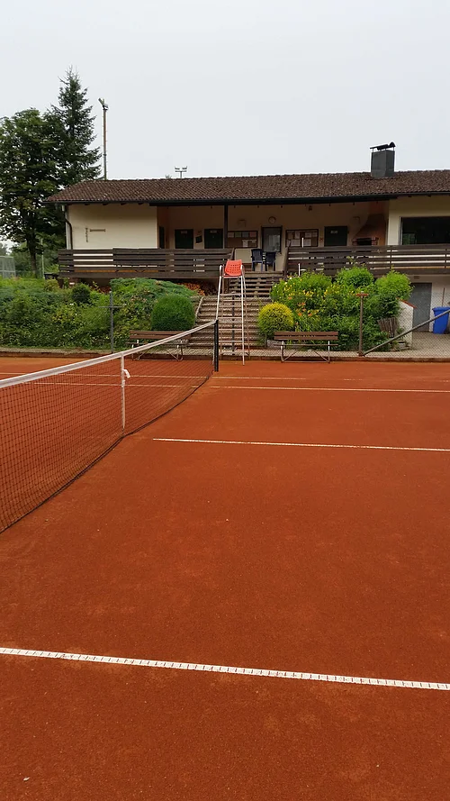 Tennisplatz mit rotem Sandbelag, einem Netz in der Mitte, dahinter ein Gebäude mit braunem Dach, einer Terrasse und Treppe, umgeben von Büschen und Bäumen.