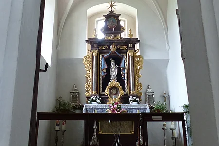 Walburgakapelle in der Stadtpfarrkirche St. Walburga Monheim Ein Blick durch eine Tür zu einem Kirchenaltar in der Walburgakapelle in Monheim, mit goldenen Verzierungen und einer Statue, der heiligen Walburga. Der Kirchenaltar ist umgeben von Kerzen und Blumen, in einem gewölbten Raum mit einer verzierten Decke.