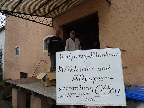 Zwei Personen stehen vor einem orangegestrichenen Gebäude neben einem Schild mit der Aufschrift "Kolping-Monheim Altkleider und Altpapier-Sammlung  Offen".