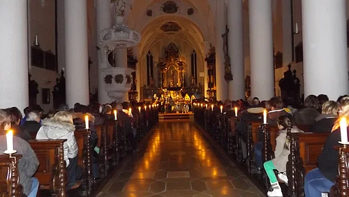 Innenraum einer Kirche mit Kerzen beleuchtet, Menschen sitzen auf Holzbänken, Blick auf den Altar im Hintergrund.