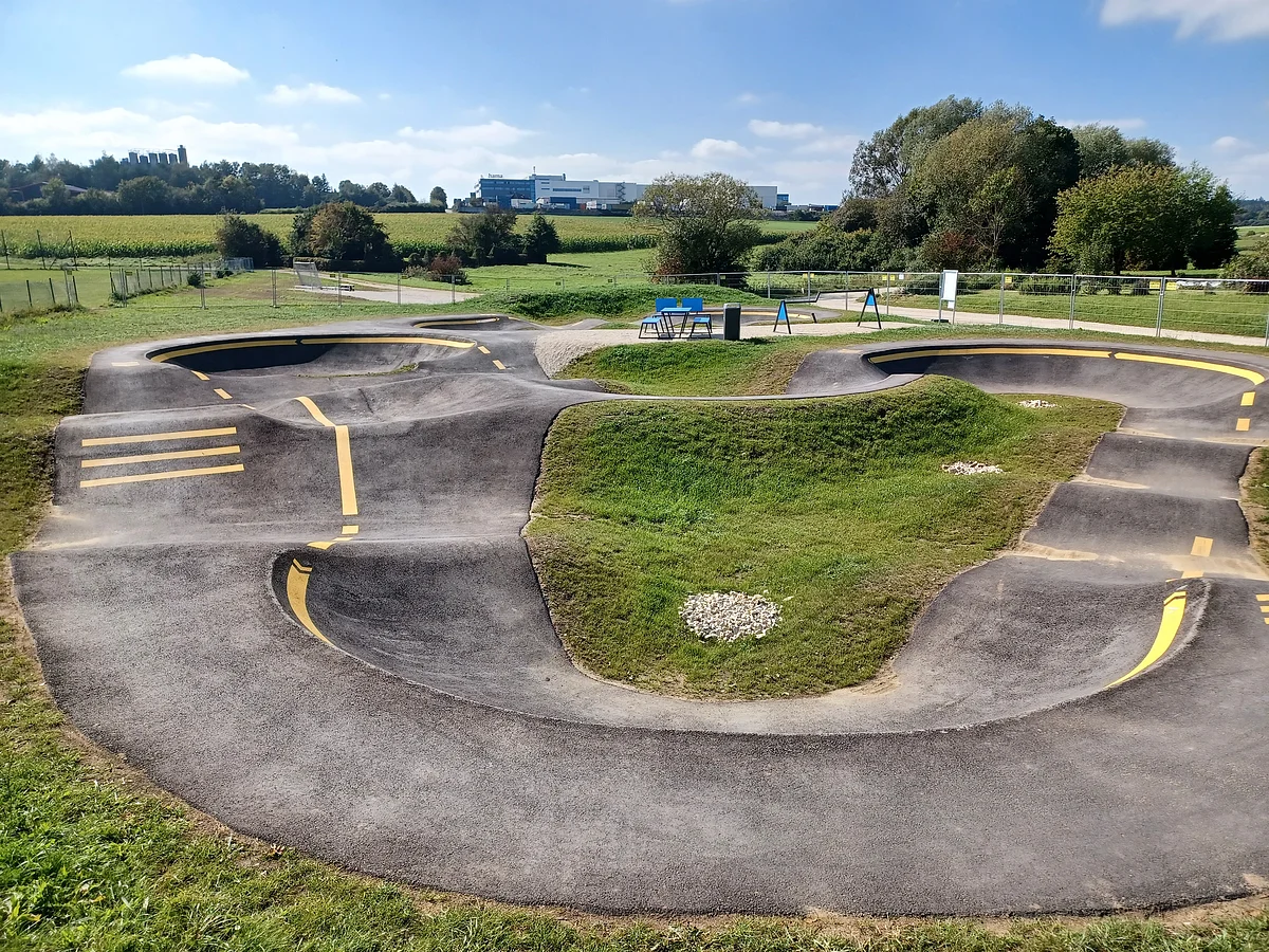 Asphaltierter Pumptrack mit gelben Markierungen in der Mitte, umgeben von grüner Landschaft und Bäumen im Hintergrund.