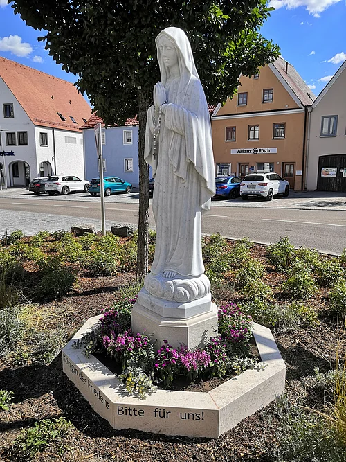 Statue einer betenden Frau mit Rosenkranz, die Marienstatue auf einem kleinen Podest, umgeben von Blumen, im Hintergrund eine Straße und ein Parkplatz mit Gebäuden.
