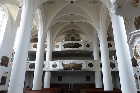 Innenansicht einer Kirche, der Stadtpfarrkirche St. Walburga in Monheim, mit hohen weißen Säulen, Gewölbedecke, Emporen mit Verzierungen und Holzbänken.