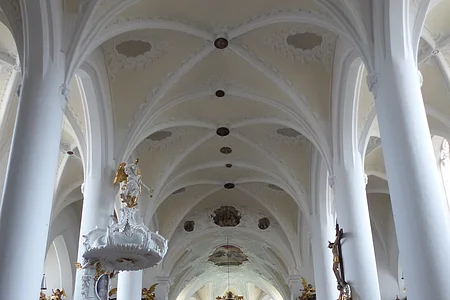 Innenansicht einer Kirche, der Stadtpfarrkirche St. Walburga in Monheim, mit hohen weißen Säulen, Gewölbedecke, Emporen mit Verzierungen und hölzernen Kirchenbänken auf beiden Seiten. Ein Altar im Hintergrund.