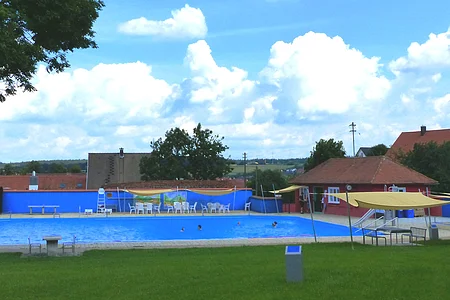Freibad mit blauem Wasser, umgeben von Rasen, Bäumen und Gebäuden unter einem bewölkten Himmel. Vorderhalb vom Becken sind Sitzmöglichkeiten.