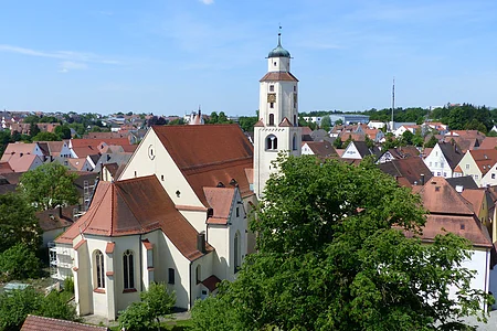 Luftbild der Stadtpfarrkirche mit Turm in einer Stadtlandschaft, umgeben von Häusern mit roten Dächern und Bäumen. Im Hintergrund blauer Himmel.