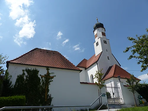 Weiße Kirche mit rotem Ziegeldach und Zwiebelturm vor blauem Himmel. Im Vordergrund ein Tor und Pflanzen.