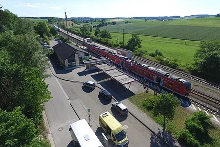 Bahnhof Otting-Weilheim Luftaufnahme eines Bahnhofs mit rotem Zug, umgeben von Feldern und Bäumen. Stehende Autos und Busse sind zu sehen.