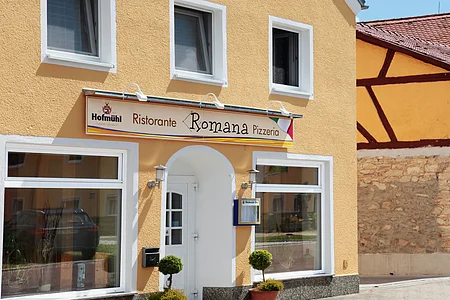 Gelbes Gebäude mit mehreren Fenstern und einem Schild "Ristorante Romana Pizzeria" über dem Eingang. Zwei kleine Bäume links und rechts vor der Tür.