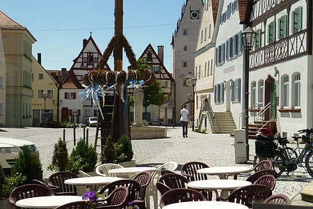 Ein Marktplatz mit Tischen und Stühlen eines Cafés im Vordergrund, Fachwerkhäusern im Hintergrund und einem Maibaum in der Mitte des Marktplatzes. Eine Person läuft auf dem Markplatz.