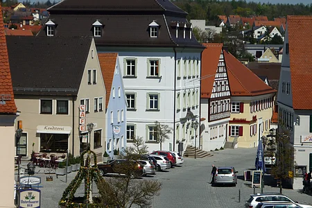 Ansicht einer gepflasterten Innenstadt mit historischen Gebäuden, geparkten Autos und einem geschmückten Oster-Brunnen. Im Hintergrund sind Wohnhäuser zu sehen.